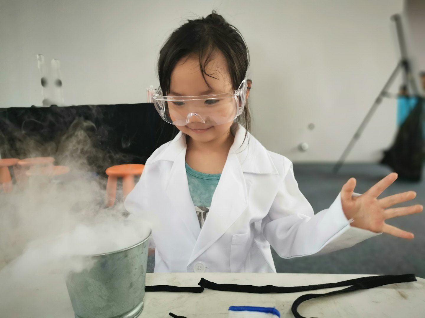 Niño con bata de laboratorio y gafas sonríe al vapor de un experimento científico.