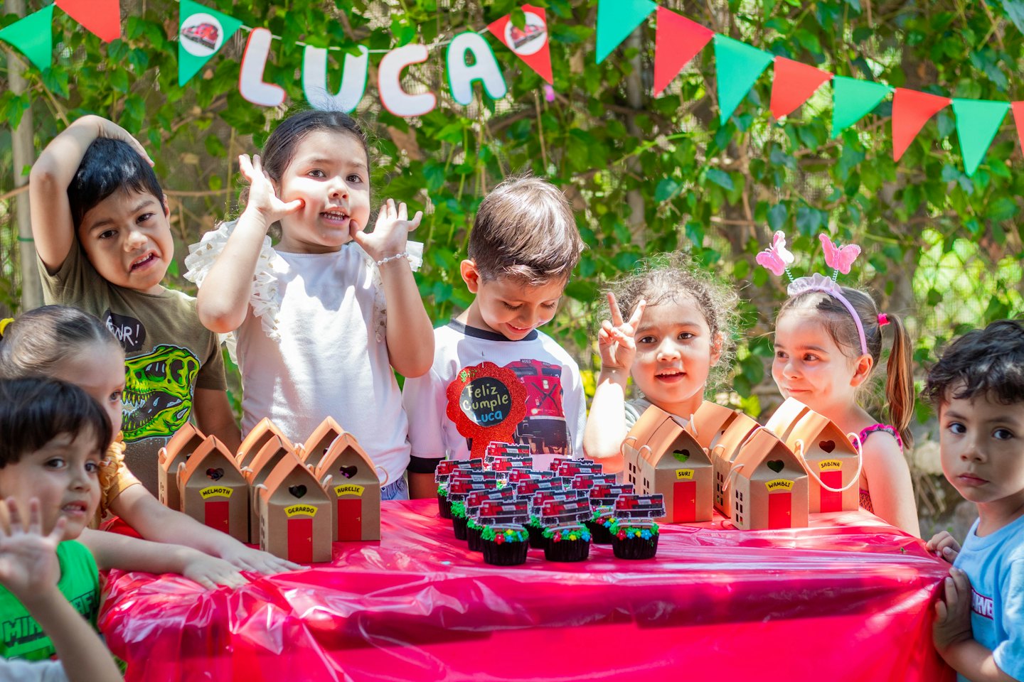 Niños celebrando un cumpleaños con pastelitos en una mesa roja.